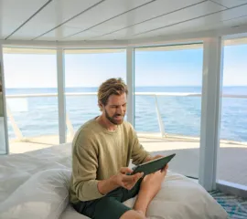 Ein Mann in einer Kabine auf einem Schiff der Mein Schiff®-Flotte. Er sitzt auf dem Bett mit Blick auf das Meer und schaut sich die neusten Aktionen von TUI Cruises auf einem Tablet an.