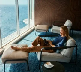 Eine blonde Frau liegt auf einer bequemen Liege in einer Lounge auf einem Schiff der Mein Schiff®-Flotte und arbeitet am Laptop mit Blick auf das Meer.