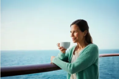 Eine brünette Frau in türkiser Strickjacke steht an der gläsernen Reling eines Schiffes der Mein Schiff-Flotte und genießt eine Tasse Kaffee beim Blick auf das Meer.