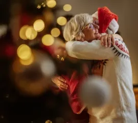 Ein Pärchen in weihnachtlichen Pullovern und Mütze umarmt sich vor dem geschmückten Weihnachtsbaum.