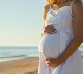 Eine schwangere Frau in einem weißen Kleid hält ihren Bauch fest, während sie am Strand steht.