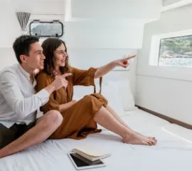 Ein Pärchen sitzt auf einem Bett in einer Kreuzfahrtkabine und erfreut sich am Ausblick. Sie haben eine Stornokabine gebucht.