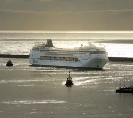 Die MSC Armonia in Seitenansicht bei der Hafeneinfahrt. Im Hintergrund geht sie Sonne unter und das Kreuzfahrtschiff wird von kleineren Booten begleitet.