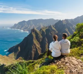 Ein Pärchen sitzt am Rande einer Klippe und schaut auf die felsige Landschaft, die grün bewachsen ist. Im Hintergrund glitzert das Meer.