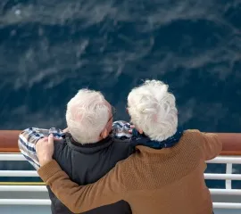 Zwei Senioren mit weißen, kurzen Haaren lehnen sich gegen die Reling und schauen vom Kreuzfahrtschiff auf das dunkle Meer.