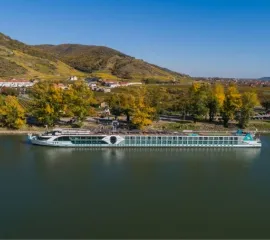 Ein Flusskreuzfahrtschiff von Phoenix liegt am Ufer des Rheins, im Hintergrund Berglandschaften.