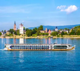 Flusskreuzfahrtschiff der Reederei 1A Vista fährt auf dem Rhein vor der Stadtkulisse von Rüdesheim mit der Brömserburg und Weinbergen unter blauem Himmel.