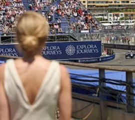 Eine elegante Frau steht an der Rennstrecke des FORMULA 1 GRAND PRIX DE MONACO 2026 und schaut sich die Formel 1 Rennautos an. Das Logo von Explora Journeys ist auf einem Werbebanner.