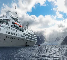 Ein Silversea Expeditionskreuzfahrtschiff ankert vor einer bergigen Gletscherkulisse in einer Polarregion. Passagiere steigen über eine Seitenpforte in schwarze Zodiac-Schlauchboote um.