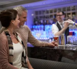 Ein Paar sitzt an der Bar im The Haven Bereich auf der Norwegian Bliss und lässt sich vom Barkeeper Drinks mischen.
