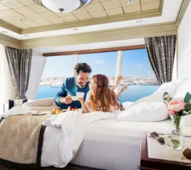 Frau und Mann liegen auf dem Bett und genießen einen kaffee in ihrer Kabine mit großem Panoramafenster und Blick auf das Meer.