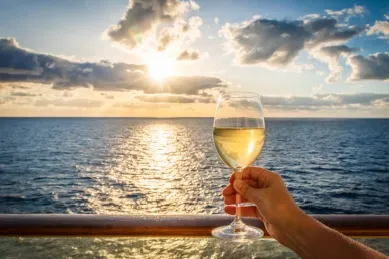 Eine Hand hält ein Glas Weißwein an der Reling eines Kreuzfahrtschiffes. Von einem leicht bewölkten Himmel aus glänzt die Sonne im Wein und wird auch vom tiefblauen Wasser reflektiert.