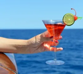Ein kühler Drink mit einer Limettenscheibe in einem Martiniglas. Eine Frau steht an Bord und hält das Glas über die Reling.