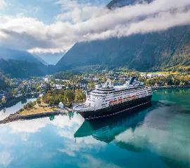 Das klassische Kreuzfahrtschiff VASCO DA GAMA von nicko cruises in einem spiegelglatten norwegischen Fjord, umgeben von majestätischen grünen Bergen und einem idyllischen Küstenort unter weichem Sonnenlicht.