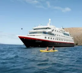 MS Santa Cruz II vor einer Küste. Zwei Personen fahren auf einem Kajak vor dem Schiff.