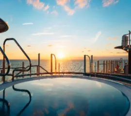 Goldener Sonnenuntergang über dem offenen Meer, fotografiert vom Außendeck eines AIDA Kreuzfahrtschiffes. Im Vordergrund sind moderne Reling-Elemente und die warme Lichtstimmung auf dem Pooldeck zu sehen, die Fernweh nach der Sommersaison 2028 weckt.