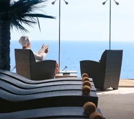 Eine Frau sitzt im Aurea Spa auf einem MSC Schiff vor einer Panorama-Scheibe. Sie trägt einen weißen Bademantel und ein weißes Handtuch auf dem Kopf und liest ein Buch. Neben Ihr steht eine Palme, im Vordergrund stehen einige Liegen..
