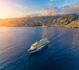 Ein Kreuzfhartschiff, dass gerade von einer sonnenbeschienen Küste ablegt und in See sticht.