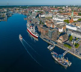 Luftaufnahme der Kieler Förde und der Hörn mit einer fahrenden Fördefähre, einem historischen Schiff am Kai und der Kieler Innenstadt im strahlenden Sonnenlicht.