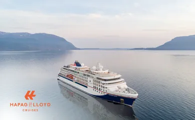 Die HANSEATIC nature von Hapag-Lloyd Cruises liegt bei diesigem Licht vor bewaldeten Bergen im Fjord.