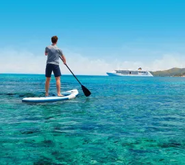 Ein Mann steht auf einem  Stand-Up-Paddle und schaut auf ein Schiff von Hapag-Lloyd Cruises. Er reist allein.