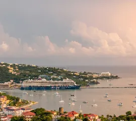 Das Kreuzfahrtschiff <i>Mein Schiff 3</i> von TUI Cruises ankert vor einer grünen Küste im warmen Abendlicht.