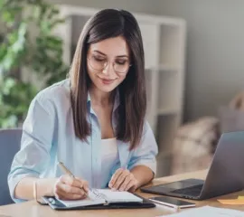 Eine Frau plant am Schreibtisch eine Reise mit nicko cruises. Ihr Laptop ist aufgeklappt und schreibt eine Notiz auf einem Block.