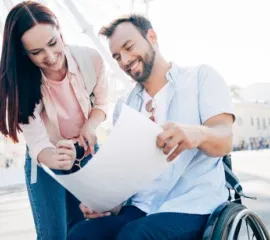 Ein  Mann im Rollstuhl und eine Frau schauen bei einem Städtetrip auf eine Papier-Karte.