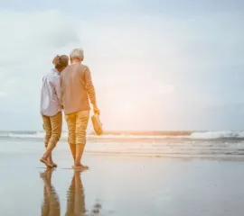 Ein älteres Pärchen läuft sorgenlos und barfuß über einen Strand und schaut sich den Sonnenuntergang an.
