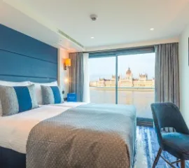 Eine Außenkabine auf der nickoVISION mit einem Doppelbett, Stuhl und Panoramafenster. Durch das Fenster ist das Budapester Parlament sichtbar.