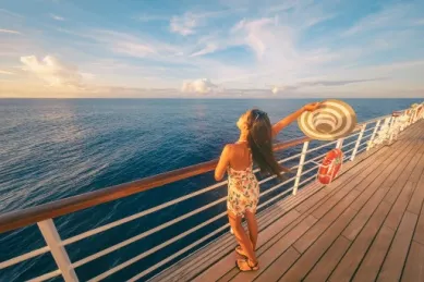 Eine Frau an der Reling eines Kreuzfahrtschiffes. Sie hält einen Sonnenhut in der ausgestreckten rechten Hand und blickt aufs Meer.