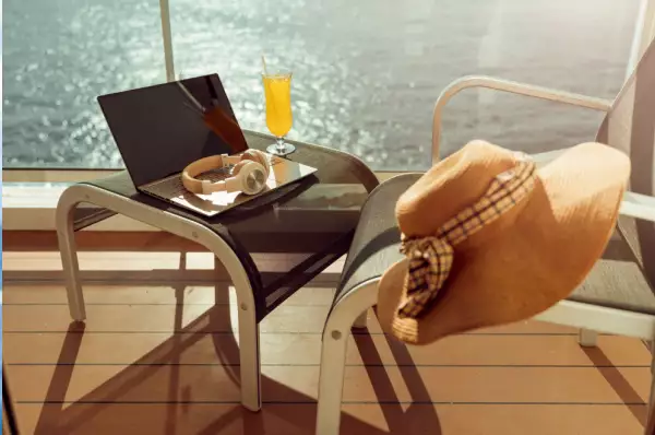 Auf dem Balkon eines Kreuzfahrtschiffs steht ein kleiner Tisch mit Laptop, Kopfhörern und Cocktail vor einem Stuhl, an dessen Lehne ein Damen-Sonnenhut hängt. Durch die Glasfront ist das Meer zu sehen.