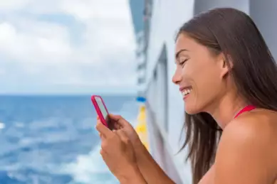 Eine junge Frau steht an der Reling eines Kreuzfahrtschiffes und schaut lächelnd auf ihr rotes Smartphone.