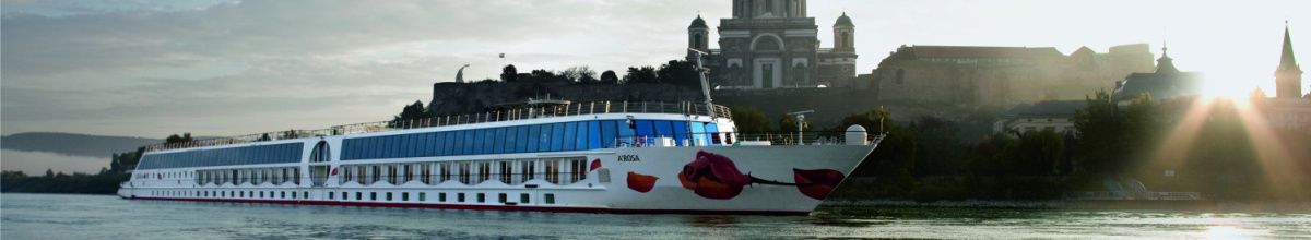 Ein A-ROSA Schiff mit dem charakteristischen Kussmund am Bug liegt auf einem Fluss, im Hintergrund die imposante Basilika von Esztergom in Ungarn bei Sonnenlicht.