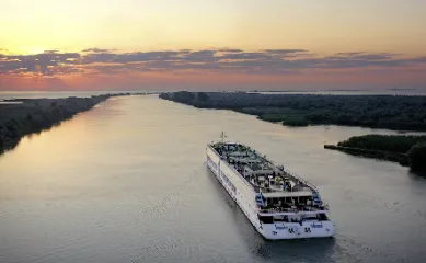A-ROSA Flusskreuzfahrt auf der Donau bei Sonnenuntergang Heckansicht eines A-ROSA Flussschiffes, das bei Sonnenuntergang einen breiten Fluss entlangfährt, umgeben von grünen Uferlandschaften.