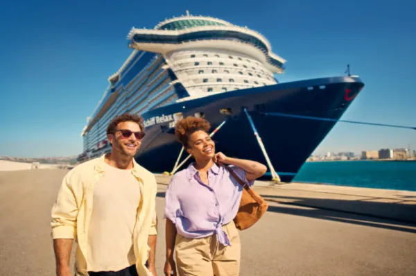Ein Mann und eine Frau spazieren lächelnd an einer Hafenpromenade entlang. Im Hintergrund liegt das große Kreuzfahrtschiff <i>Mein Schiff Relax</i> bei sonnigem Wetter am Kai.