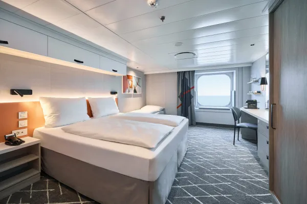 Blick in eine modern eingerichtete Außenkabine auf der <i>Mein Schiff Relax</i> mit einem großen Doppelbett, einem Schreibtisch und einem runden Fenster mit Blick auf das Meer.