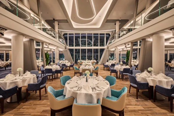 Das elegante, zweistöckige Hauptrestaurant Atlantik am Heck der <i>Mein Schiff Relax</i> bei Nacht, mit eingedeckten Tischen und großer Panorama-Fensterfront.