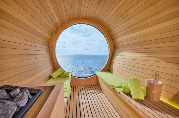 Innenansicht der hölzernen Fass-Sauna auf der <i>Mein Schiff Relax</i> mit einem großen, runden Fenster, das einen weiten Blick auf das offene Meer bietet.