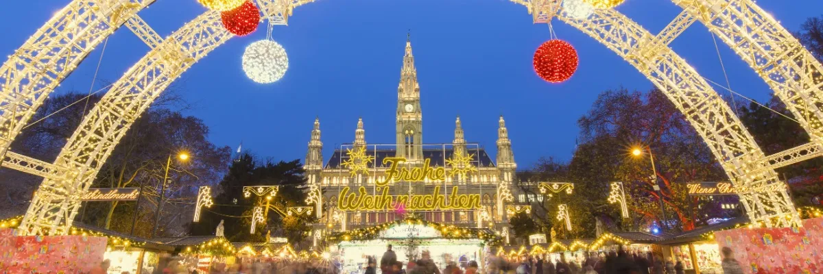 Weihnachtsmarkt vor dem Wiener Rathaus. Im Vordergrund ein Lichterbogen und eine goldene Leuchtschrift Frohe Weihnachten, dahinter die Buden des Weihnachtsmarktes