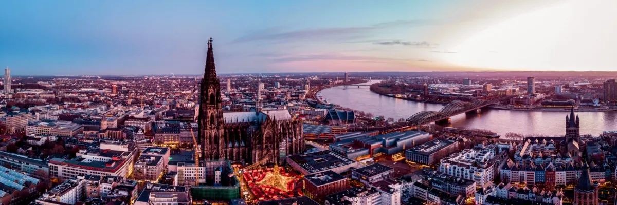 Luftbild des Kölner Doms mit davor liegendem Weihnachtsmarkt in der Abenddämmerung. Am rechten Bildrand sind außerdem der Rhein und die Hohenzollernbrücke zu sehen.
