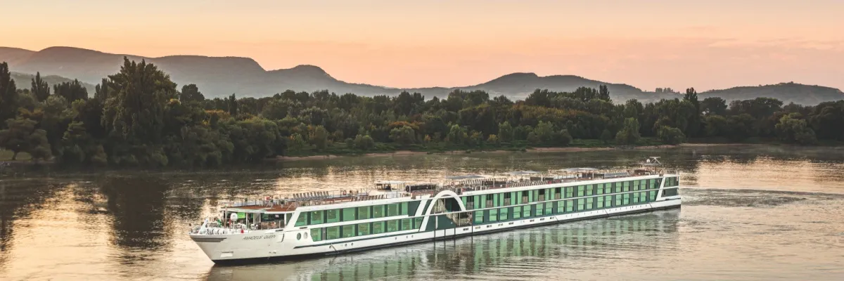 AMADEUS Queen im Sonnenuntergang auf der Donau. Im Hintergrund ein baumbestandenes Ufer sowie eine Bergkette.