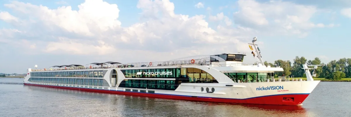 Seitenansicht von nickoVISIOn auf einem Fluss. Im Hintergrund ein grünes Ufer mit einigen Bäumen, darüber ein blauer Himmel mit dicken weißen Wolken.