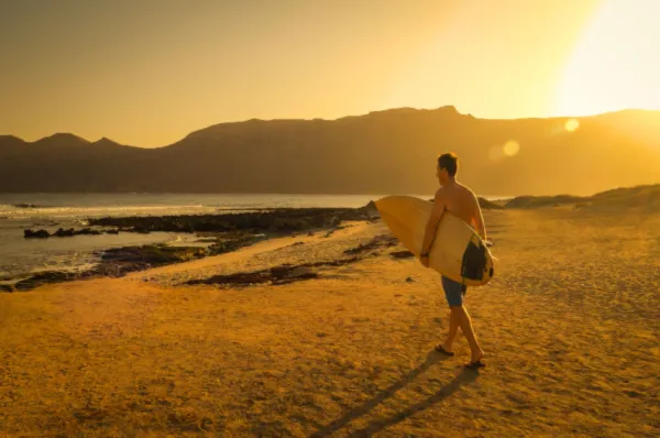 Ein Mann mit Surfbrett Ein Surfer der mitsamt Surfbrett auf das Meer zuläuft, während im Hintergrund die Sonne die Landschaft in warmes Gold taucht.