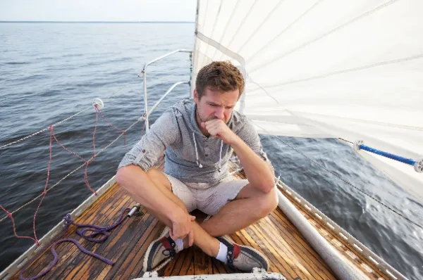 Mann sitzt mit Übelkaut auf dem Deck eines Segelbootes, Symbol für die Symptomatik bei Seekrankheit bei einer Kreuzfahrt.