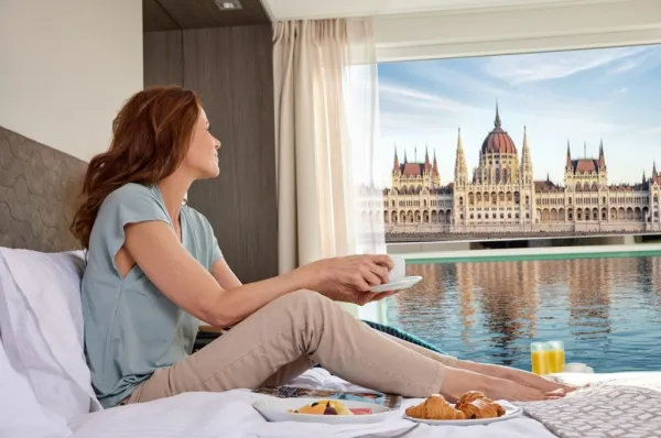 Kreuzfahrt auf der Donau Eine Frau sitzt auf dem Bett ihrer Kabine auf einer Flusskreuzfahrt mit Sicht auf das Parlament in Budapest.