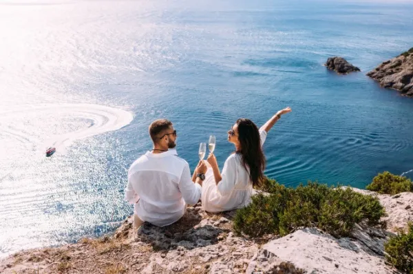 Symbolische Hochzeiten Ein Paar sitzt mit Sektgläsern auf einem Felsen an der sonnigen Küste. Sie haben gerade symbolisch geheiratet.