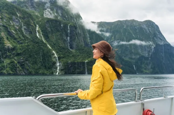Eine Frau lehnt an der Reling eines Schiffes und schaut auf eine Bergkette. Sie trägt einen gelben Regenmantel und einen Hut.