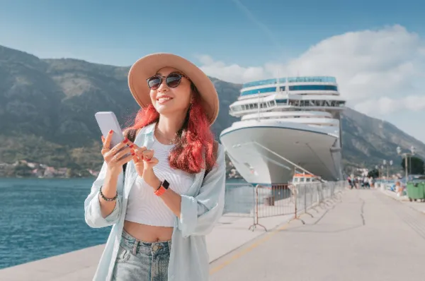 Single Kreuzfahrt: Unterwegs im Hafen und bestens informiert Lächelnde Frau mit Hut und Sonnenbrille nutzt ihr Smartphone am Kai, im Hintergrund ein großes Kreuzfahrtschiff im Hafen vor einer Bergkulisse – Planung oder Vernetzung während einer Single Kreuzfahrt.
