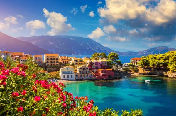 Idyllische Bucht als Ziel einer Mittelmeer-Kreuzfahrt Malerische Ansicht einer Bucht in Griechenland bei strahlendem Sonnenschein, ein Highlight jeder Mittelmeer-Kreuzfahrt. Leuchtend pinke Blumen im Vordergrund, bunte Häuser und türkisblaues Wasser schaffen eine perfekte Urlaubskulisse.